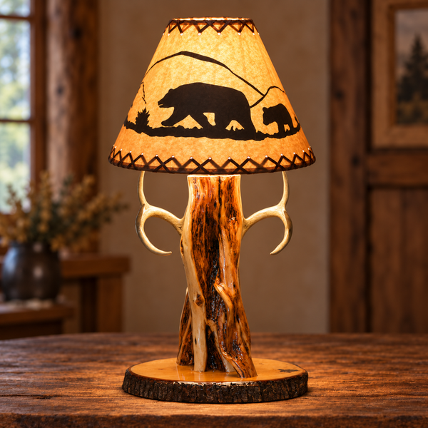 A rustic table lamp with a natural wood bark base, antler-like side supports, and a conical shade printed with a black bear and cub silhouette and geometric border details, shown on a wooden table in a warm cabin-style room.